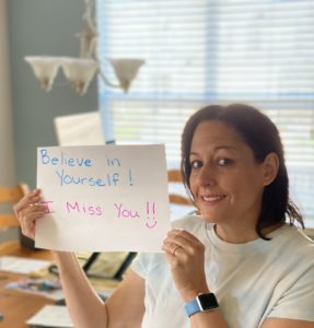 Lorraine Elias smiling at camera holding sign saying "Believe in yourself! Miss you!"