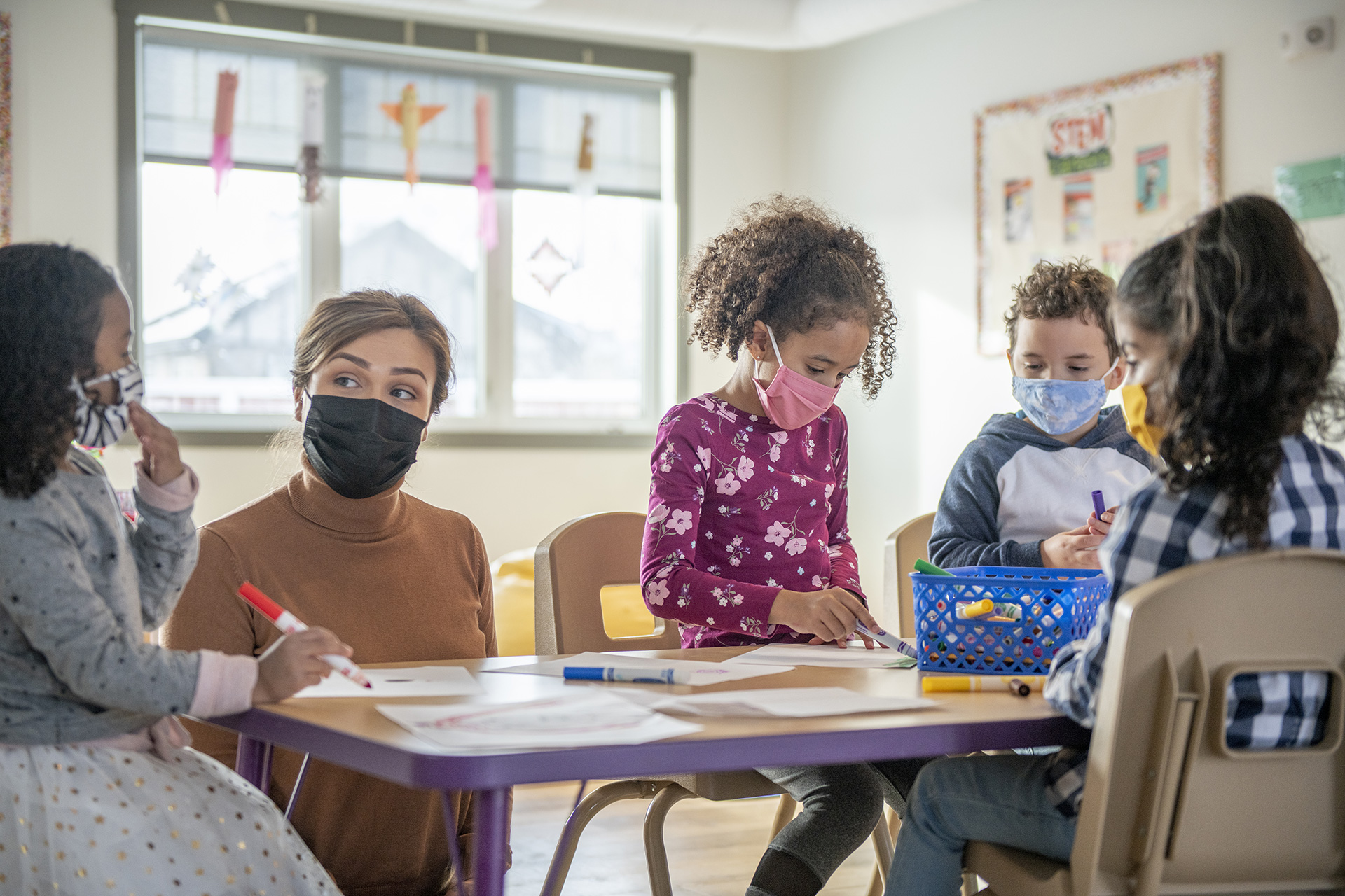 teacher in black mask working alongside children in masks in classroom